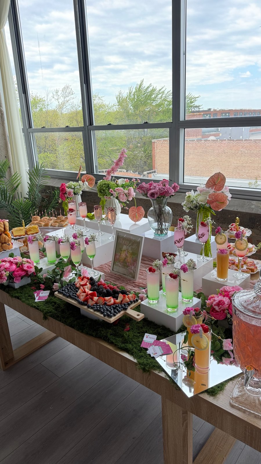Bloom & Bubbly brunch table with moss runner, floral arrangements, layered drinks, champagne flutes, and a framed Bloom & Bubbly sign by a window.