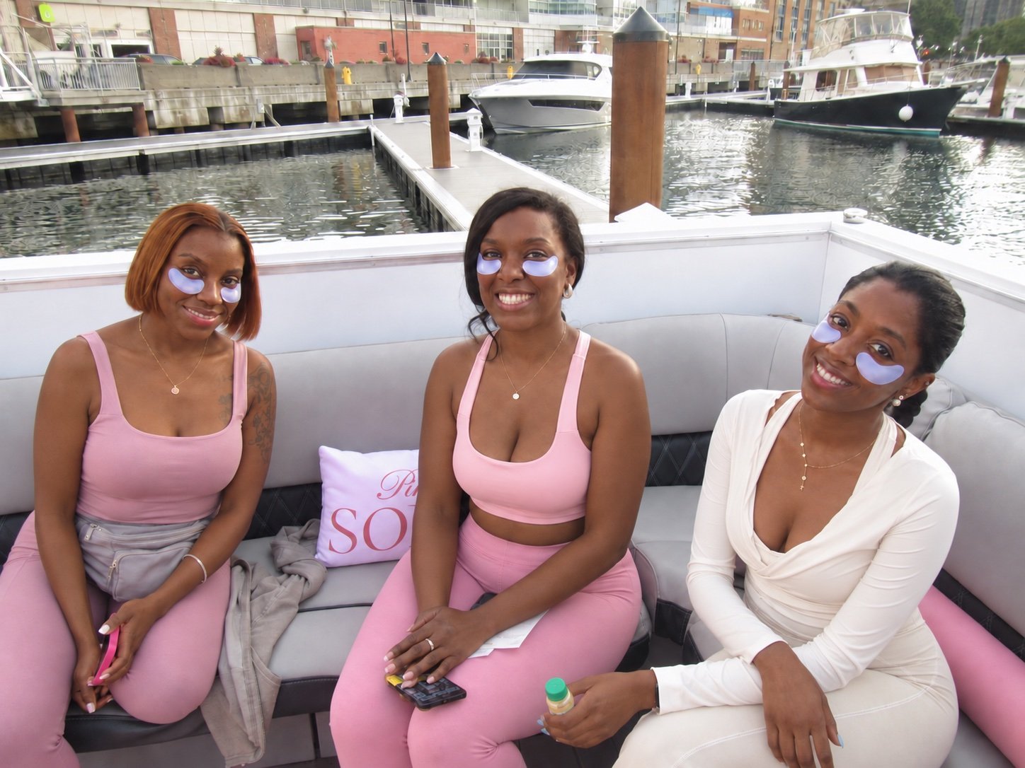 Three guests on a marina boat wear purple under-eye patches and smile together, with docks and yachts behind them.