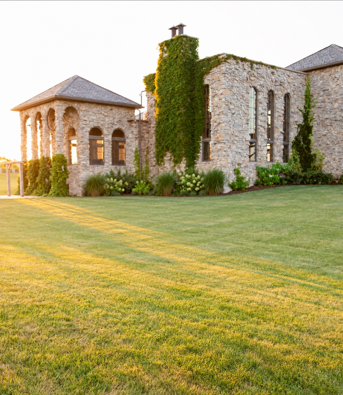 Stone château with ivy-covered tower and arched patio at golden hour across a wide green lawn.
