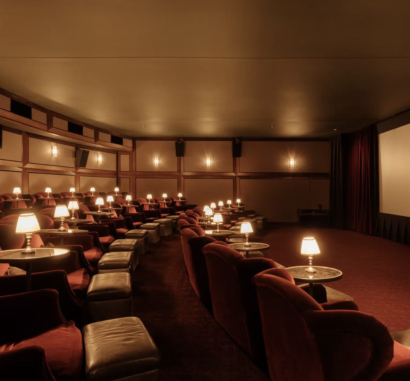 Private Soho House screening room with plush burgundy velvet armchairs, leather footstools, and warm table lamps facing a curtained cinema screen.